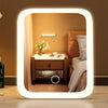 A glowing LED vanity makeup mirror with a rounded frame reflecting a warm-toned bedroom interior.