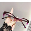 A hand holds a pair of purple cat eye reading glasses with clear frames against a light background.