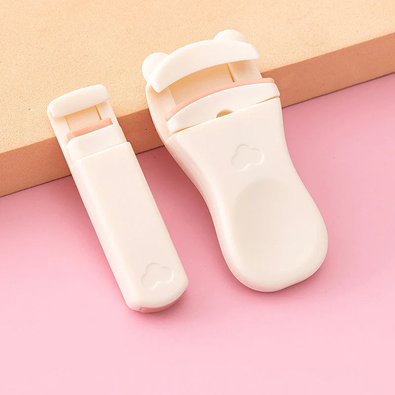 A white cartoon mini eyelash curler is shown lying on a pink surface.