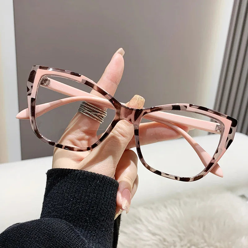 A hand holds a pair of pink and black patterned cat eye reading glasses against a light background.