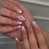 A close-up of a hand displaying 24-piece almond press-on nails featuring floral and butterfly designs.