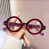 A hand holds a pair of round, pink leopard print acetate reading glasses with anti-blue light lenses.