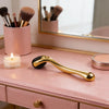 A gold-colored titanium micro derma roller tool rests on a pink vanity surface next to a candle and makeup brushes.