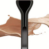 A black makeup brush with a pink hair-like synthetic fiber is shown against a background of liquid cosmetic product.