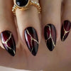 A close-up of a hand displaying dark burgundy press-on false nails with gold leaf accents and a gold ring.