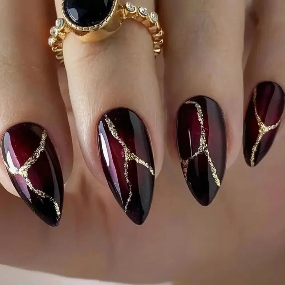 A close-up of a hand displaying dark burgundy press-on false nails with gold leaf accents and a gold ring.