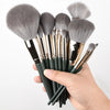 A hand holds a set of ten professional makeup brushes with silver and gray bristles.