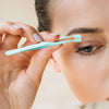 A hand holds a light blue stainless steel eyelash cluster tweezers near an eye for application.