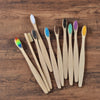 A collection of ten natural bamboo toothbrushes in various colors laid out on a wooden surface.