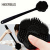 A black long-handle silicone body brush with a dual-sided bristled head being held by a hand.