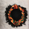 Black afro puff turban wig with an orange and green patterned fabric resting on a white fluffy surface.