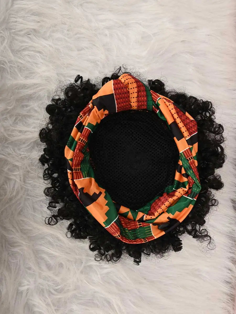 Black afro puff turban wig with an orange and green patterned fabric resting on a white fluffy surface.