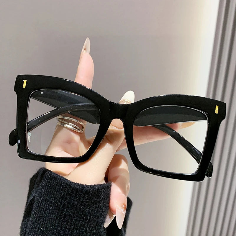A hand holds a pair of black square frame reading glasses with a silver detail against a light background.