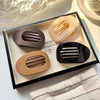A display of four oval flat hair clips in various neutral tones, including beige, brown, and black, arranged in a dark tray.