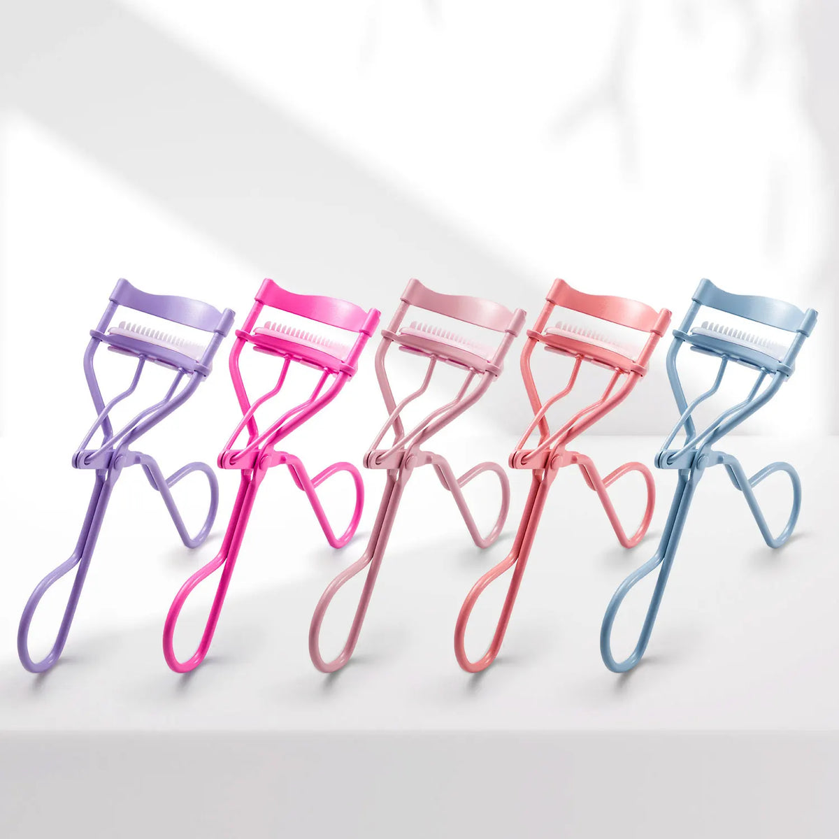 A row of five precision eyelash curlers in various pastel colors, including purple, pink, and blue, displayed on a white surf