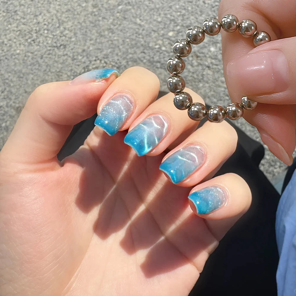 Une main montre un ensemble de aimants à ongles en argent et du vernis bleu iridescent sur les ongles.