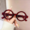 A hand holds a pair of round, reddish-brown acetate reading glasses with clear lenses against a light background.