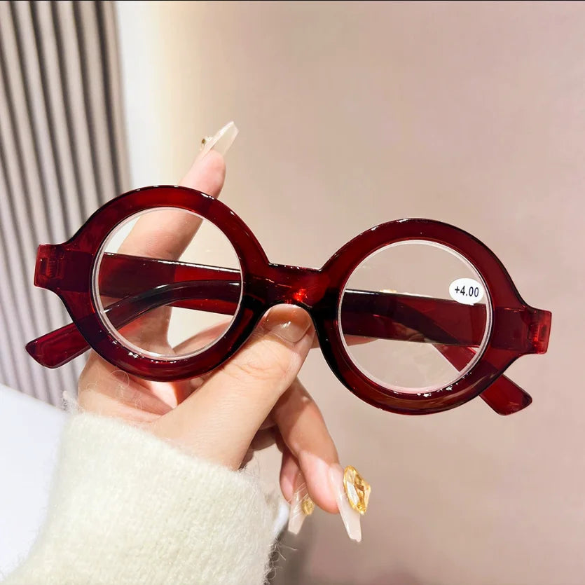 A hand holds a pair of round, reddish-brown acetate reading glasses with clear lenses against a light background.