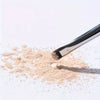 A precision eyeshadow brush is shown with light beige powder scattered on a white surface.