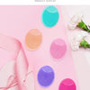 A display of various colored silicone facial cleansing brushes on a pink background.