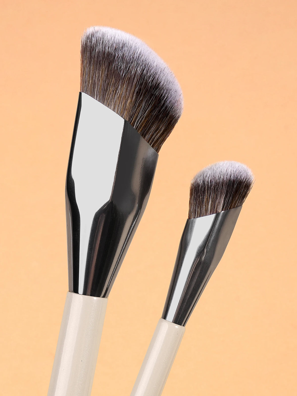 A pair of professional makeup brushes with silver ferrules and natural hair bristles against a light orange background.