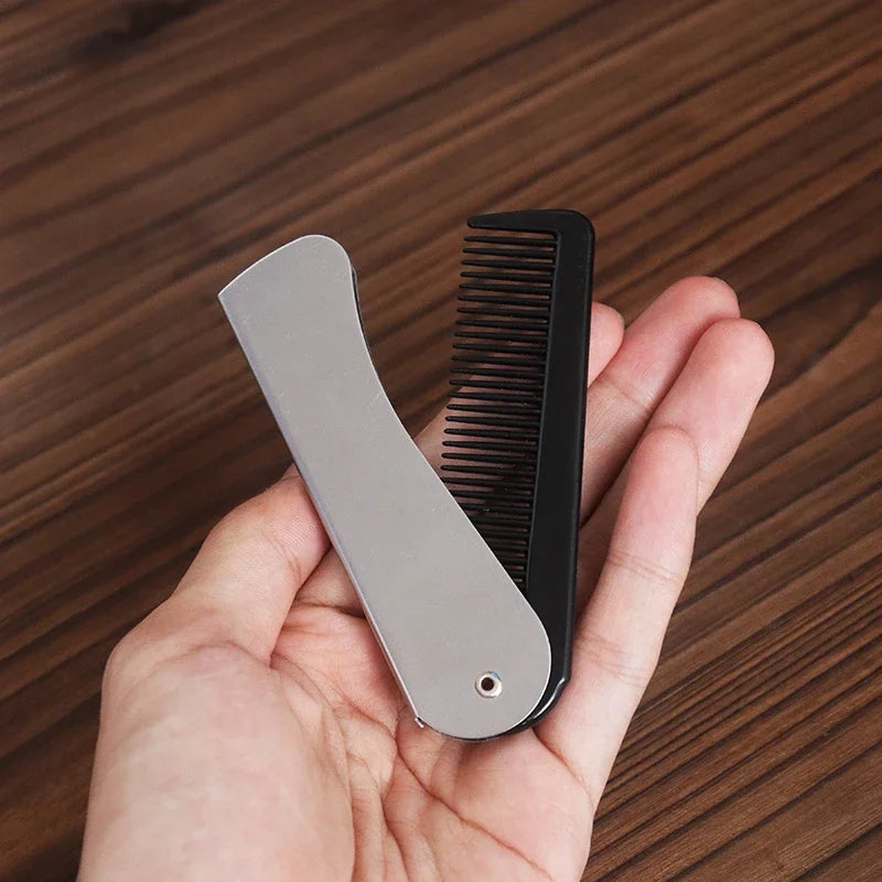 A hand holds a foldable anti-static steel comb with a silver body and black bristles against a wooden background.