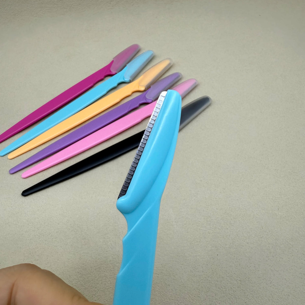Une main tient une collection de ciseaux de précision pour sourcils de différentes couleurs, y compris rose, jaune et noir.
