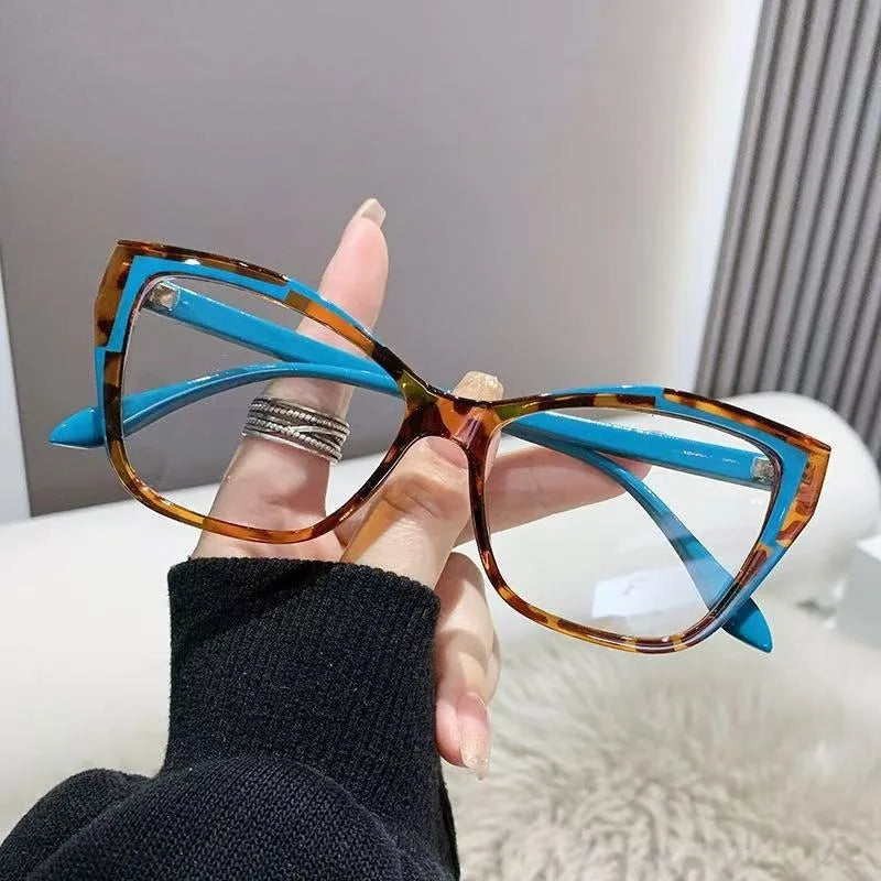 Une main tient une paire de lunettes à monture chat-œil teintées d'orange avec des accents bleus sur fond blanc.
