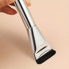 A hand holds a silver oval foundation brush with a black synthetic hair tip.