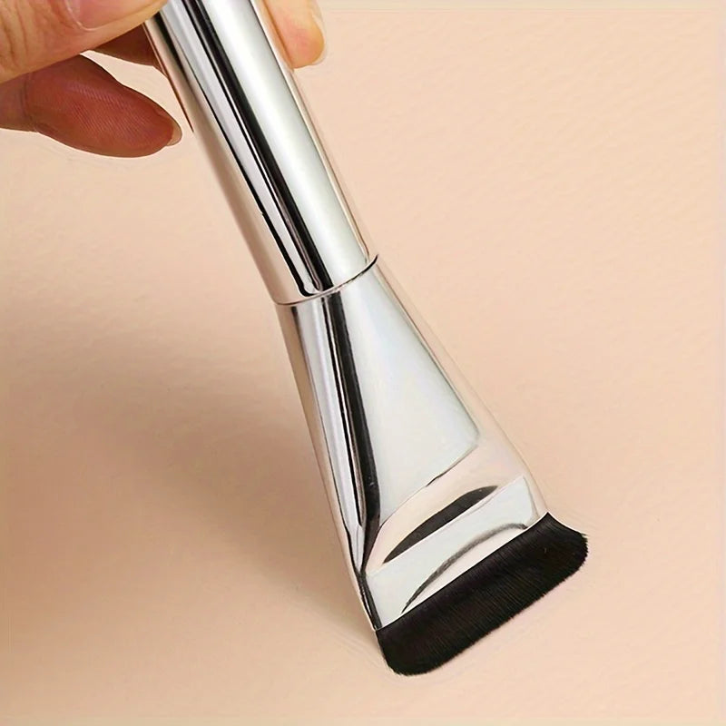 A hand holds a silver oval foundation brush with a black synthetic hair tip.