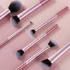 A set of seven professional makeup brushes with pink and rose gold handles and black and grey bristles displayed on a light b