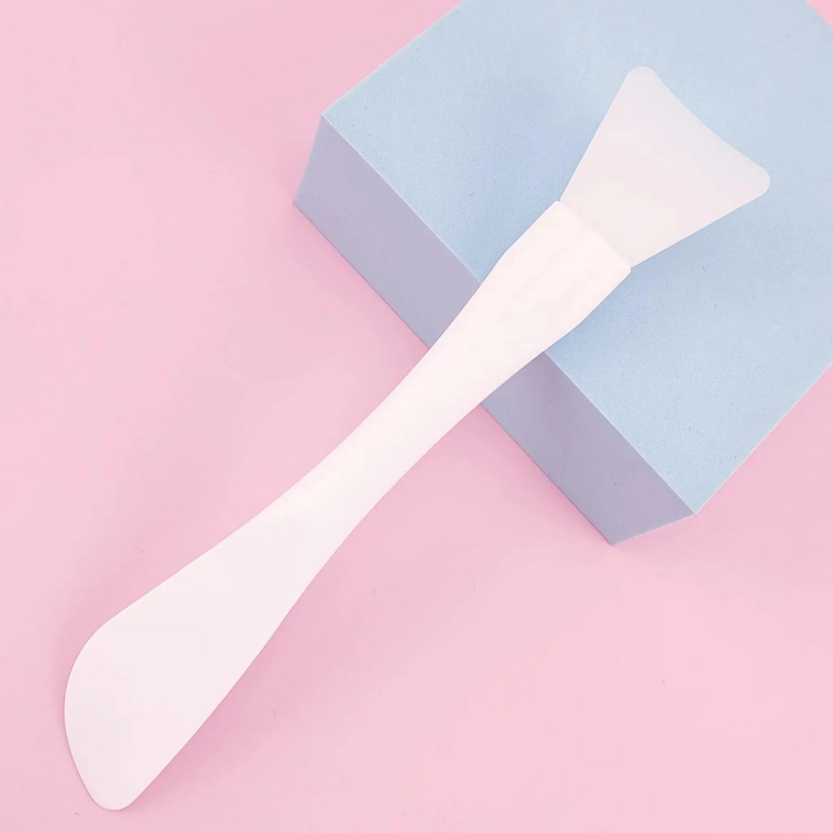 A white silicone facial mask brush with an integrated scraper resting on a light blue square box.