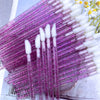 A collection of purple crystal lip brushes and white lash spoolies arranged diagonally on a surface.
