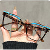Una mano sostiene unas gafas de lectura marrones tortugas con detalles azules contra un fondo gris.