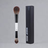 A black makeup brush with a white band and a brown hair, next to its black packaging box.