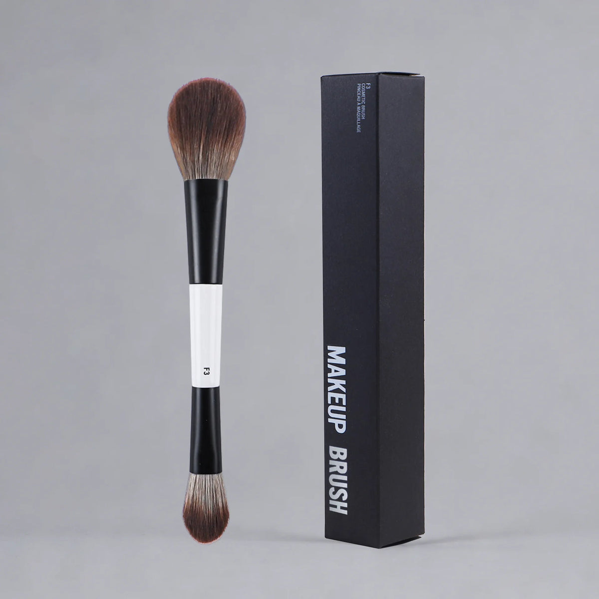 A black makeup brush with a white band and a brown hair, next to its black packaging box.