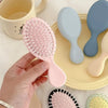 يد تمسك بفرشاة تصفيف شعر وردية صغيرة ومساج بجانب فرش سيليكون ملونة على طبق أصفر.