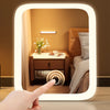 A hand is touching the frame of an LED vanity makeup mirror with adjustable light, showing a bedroom interior.