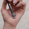 A hand holds a precision concealer brush with a silver ferrule and soft, fluffy bristles.