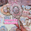 A hand holds a decorative makeup mirror set featuring pink and silver accents on a light background.