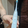 A hand holds a light blue electric toothbrush with a blue brush head and white cleaning indicators.