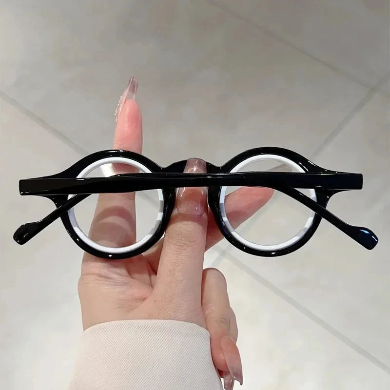 Une main tient une paire de lunettes de lecture à monture noire et verres clairs sur un fond clair.