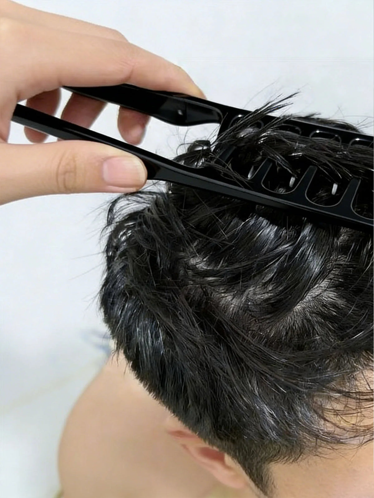 A hand is using a black hair styling comb set to style dark hair on a person's head.