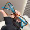 A hand holds a pair of orange-tinted cat eye reading glasses with bright blue frames against a dark sweater.