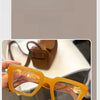 Une paire de lunettes de lecture carrées rétro, teintées en jaune avec des montures orange, tenues par une main.