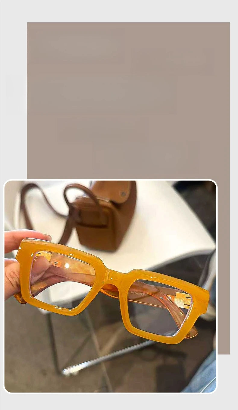 Une paire de lunettes de lecture carrées rétro, teintées en jaune avec des montures orange, tenues par une main.