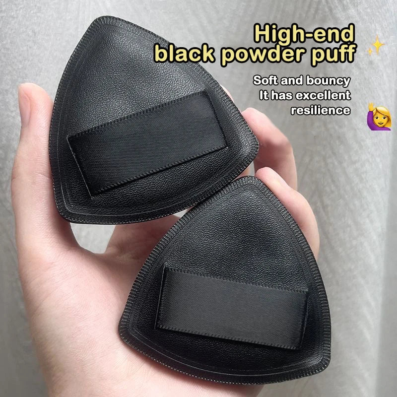 A hand holds a black powder puff brush with a triangular shape, demonstrating its soft and bouncy texture.
