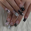 A close-up of a hand displaying gothic Y2K press-on nails featuring black and white designs with cross and bow motifs.