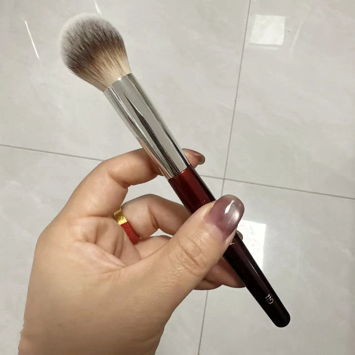 Une main tient un pinceau de maquillage liquide angulé avec un manche brun et une ferule argentée sur un fond de carreaux cla