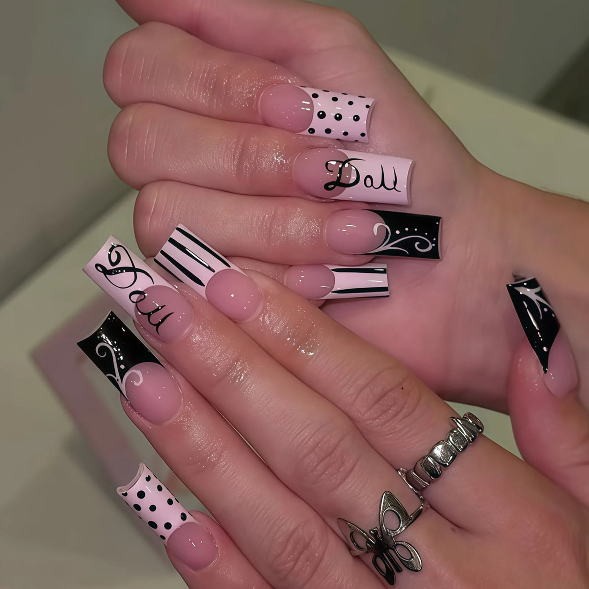 A hand displays gothic Y2K press-on nails featuring black and white designs with cross and bow motifs.